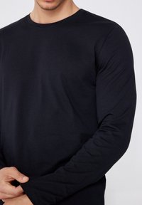 Schwarzes langärmliges T-Shirt aus weichem Baumwollmaterial, klassischem Rundhalsausschnitt, figurbetonter Passform, glatter Textur und dezenten Stichdetails an den Ärmeln.