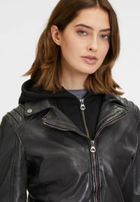 Giubbotto in pelle nera da moto con zip frontale, colletto a scatto e dettagli sulle spalle, indossato sopra una felpa nera con cappuccio.