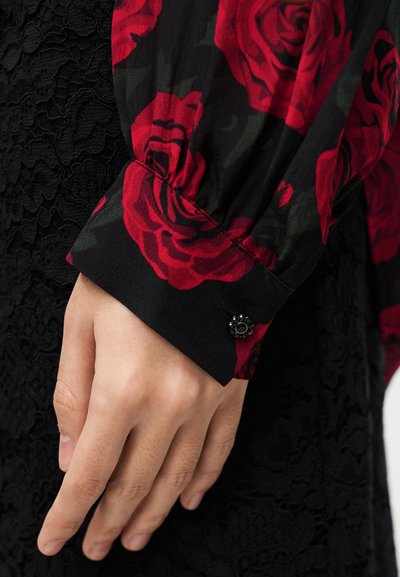 Μαύρο φλοράλ μπλουζάκι με μοτίβα κόκκινων τριαντάφυλλων, με μαύρο υφασμάτινο δαντελένιο φόντο. Η μανσέτα έχει διακοσμητικό μαύρο κουμπί.