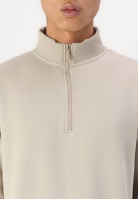 Béžová mikina na zip s vysokým límcem, vyrobená z hladké tkaniny. Disponuje krátkým zipem na krku a štíhlým designem.