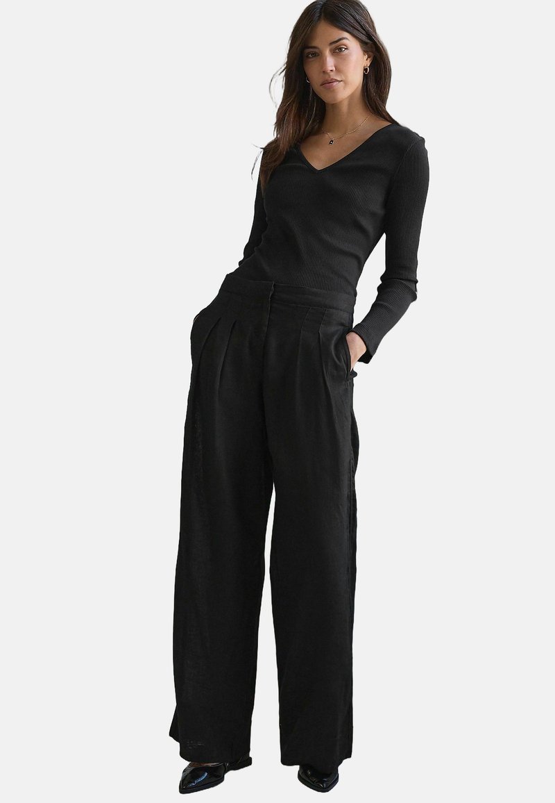 Top lungo a maniche ribbed nere con scollatura a V abbinato a pantaloni neri a gamba larga con pinces e tasche. Tessuto liscio e design minimale.
