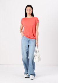 Korallfarget kortermet skjorte, avslappet passform, kombinert med lyseblå vid jeans. Modellen holder en liten hvit håndveske og står mot en ensfarget bakgrunn.