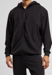 Sudadera negra con cremallera y textura suave, que incluye una capucha, dos bolsillos frontales y detalles de costura sutiles. Combinada con pantalones a juego.