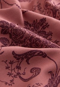 Weicher staubig rosa Stoff mit dunkelvioletten Blumen- und Ornamentmustern, in Nahaufnahme mit natürlichen Falten und Texturdetails gezeigt.