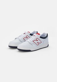 Białe sportowe sneakersy z granatowymi akcentami, wykonane z materiału przypominającego skórę, z perforowanym przodem, czerwonym logo na boku i teksturowaną gumową podeszwą.