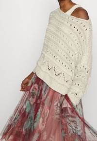 Pull crème en maille avec motif ajouré, design à épaules dénudées ; superposé sur une jupe en tulle bordeaux à imprimé floral transparent.