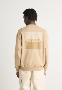 Beige sweatshirt with a large light brown square and the word "HUGO" printed in cream. Features ribbed cuffs and hem, with a relaxed fit.