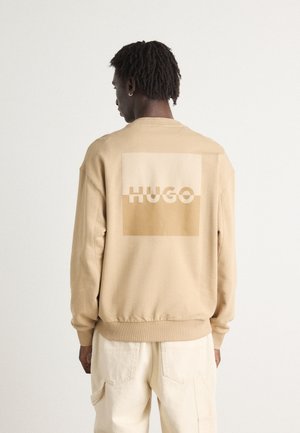 Beige sweatshirt with a large light brown square and the word "HUGO" printed in cream. Features ribbed cuffs and hem, with a relaxed fit.