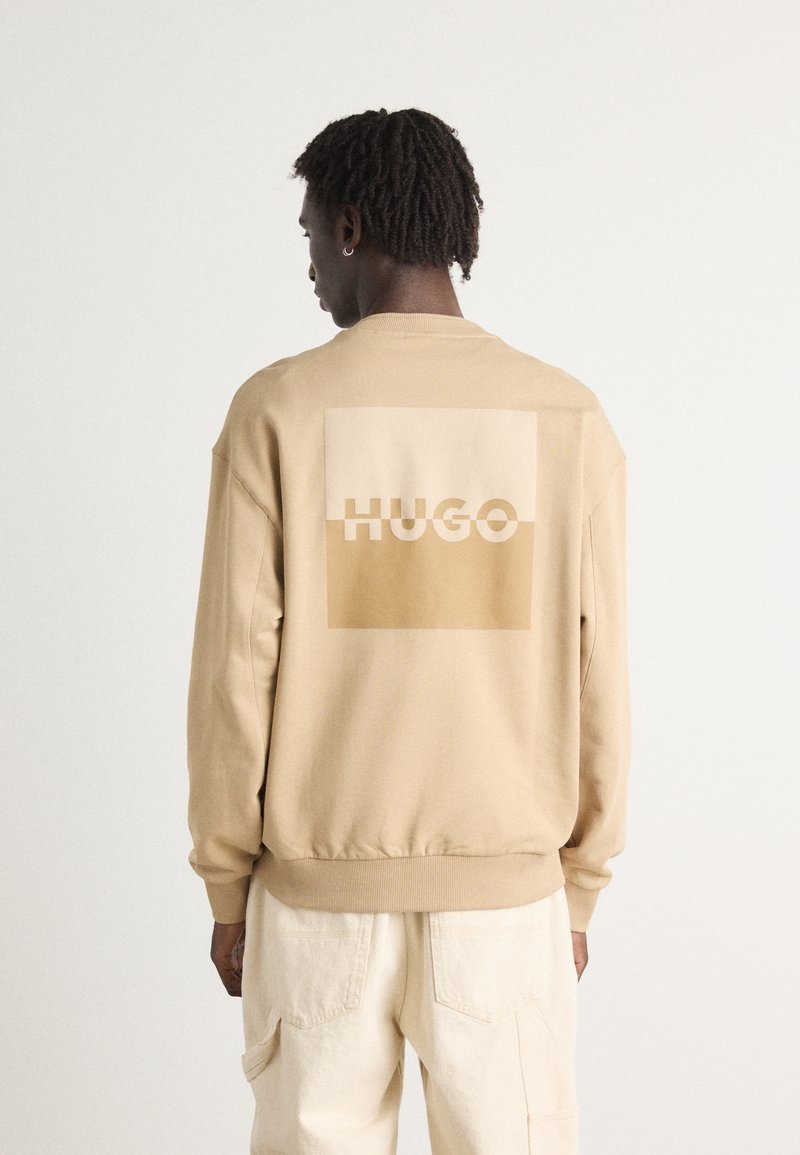 Beige sweatshirt with a large light brown square and the word "HUGO" printed in cream. Features ribbed cuffs and hem, with a relaxed fit.