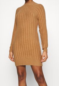 Abito in maglia color cammello, caratterizzato da una trama a costine, maniche lunghe e un collo alto. Design avvolgente con un orlo leggermente svasato.