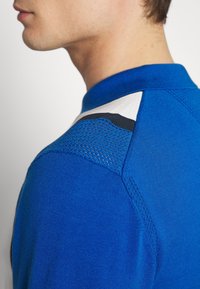 Modrý pletený svetr s texturovaným, prodyšným síťovaným detailem na rameni. Má jednobarevný vzhled a klasický límec.