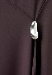 Ciemnofioletowa tkanina o gładkiej fakturze, z srebrnym, łezkowatym akcentem przy zapięciu, podkreślającym unikalne detale wzornicze.