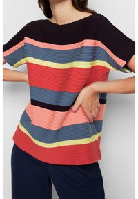 Vrouw in een multikleurige gestreepte korte mouwen gebreid top met marineblauwe broek, poseert met handen op de heupen tegen een effen achtergrond.