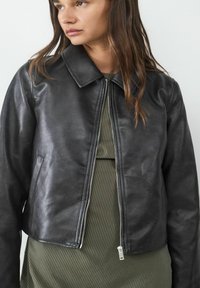 Giubbotto di pelle nera con colletto e chiusura zippata. Dotato di tasche laterali e design accorciato, indossato sopra un vestito a righe verdi.