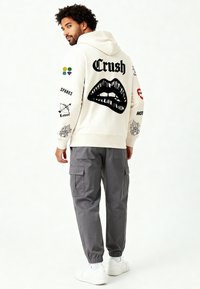 Felpa con cappuccio crema con labbra grafiche nere e testo "Crush"; toppe colorate sulle maniche. Abbinata a pantaloni cargo grigi e scarpe bianche.