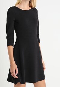 Svart ribbad klänning med rund halsringning, trekvartsärmar och en utsvängd kjol. Tygtexturen är strukturerad och något elastisk.