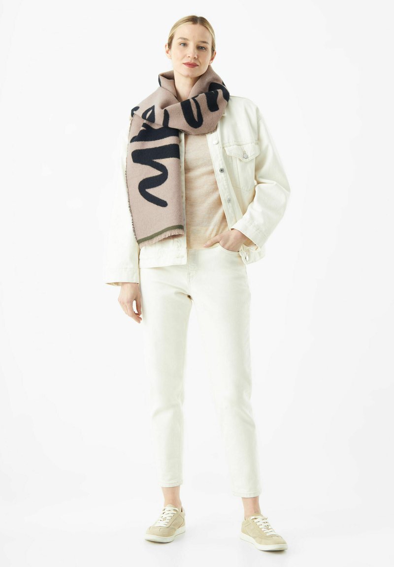 Beige wollen sjaal met donkere letters gedrapeerd rond de nek, gecombineerd met een lichte trui en een wit jack en broek, afgewerkt met beige sneakers.