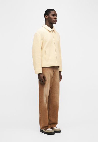 Άνδρας που φοράει εκρού ζα κέτα με φερμουάρ, καφέ παντελόνι με φαρδιά μπατζάκια και δίχρωμα loafers, στέκεται πλάγια μπροστά σε απλό φόντο.