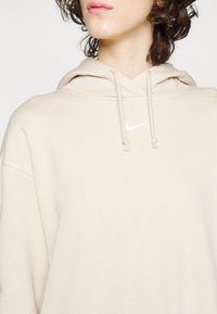 Sweat à capuche beige en tissu doux. Il comprend une poche avant, une capuche avec cordon de serrage et un logo Nike blanc sur la poitrine.