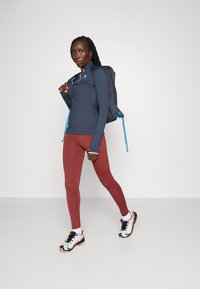Donkerblauwe langemouwen top, koraalkleurige accenten, kastanjebruine leggings, en zwarte sneakers met witte en oranje details; met een donkere rugzak met blauwe bandjes.