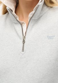 Sudadera gris claro con cuello con cremallera y forro interior blanco. Presenta un pequeño logo azul de 'Superdry' y tejido texturizado.
