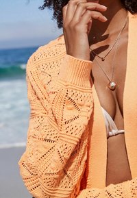 Pomarańczowy sweter na drutach z otwartym przodem i wzorzystą fakturą, noszony na jasnym topie bikini, na tle plaży.