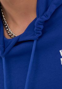 Modrá mikina s kapucí a šňůrkou, s žebrovanou strukturou, na výstřihu je viditelný stříbrný řetízek. Na látce je bílý text.