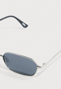 Γυαλιά ηλίου με ασημένιο μεταλλικό πλαίσιο, φακούς με γκρι απόχρωση και κομψό, επιμηκυμένο οβάλ σχήμα. Μαύρες άκρα σκελετού με λεπτομέρεια λογότυπου.