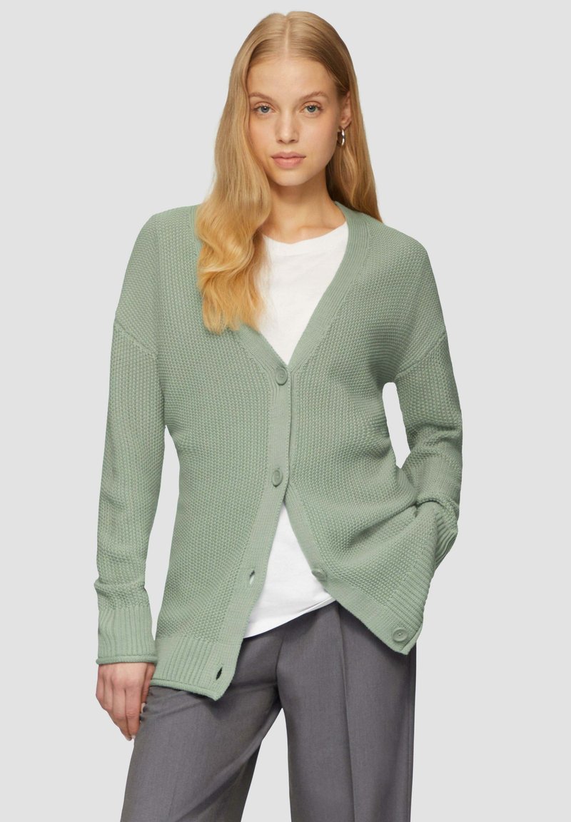 Hellgrüner Strickcardigan mit V-Ausschnitt und Knopfverschluss. Verfügt über ein strukturiertes Material, lange Ärmel und gerippte Bündchen. Wird über einem weißen Hemd getragen.