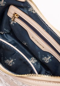 Beige Reißverschlusstasche mit goldenen Beschlägen, gefüttert mit marineblauem Stoff, der ein sich wiederholendes helles Logomuster zeigt. Enthält eine kleine Innentasche.