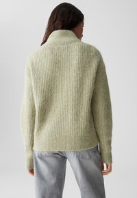 Hellgrüner, strukturierter, gerippter Pullover mit hohem Kragen und langen Ärmeln, kombiniert mit hellgrauen Jeans.