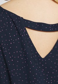 Mörkblå blus med små rosa prickar, har en V-formad ryggöppning och ett band över axlarna, tillverkad av lättviktsmaterial.