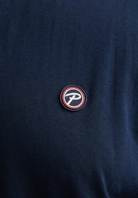 Marineblaue Kurzarmbluse mit einem runden, gestickten Aufnäher, der ein schwarz-weißes Logo zeigt, umgeben von roten und schwarzen Rändern.