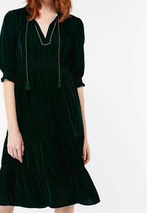 Femme portant une robe en velours vert foncé longueur genoux avec manches bouffantes, taille froncée, jupe à volants et détail de cordon à franges au niveau du col.