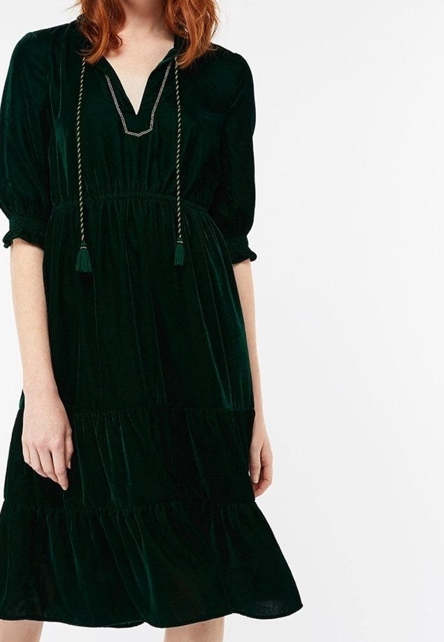 Femme portant une robe en velours vert foncé longueur genoux avec manches bouffantes, taille froncée, jupe à volants et détail de cordon à franges au niveau du col.