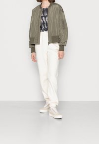 Bomber jacket verde oliva, maglietta tie-dye, pantaloni felpati crema con logo e sneakers beige con suola bianca. Materiali morbidi, vestibilità rilassata.