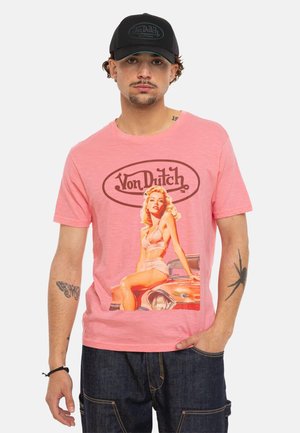 Homme portant une casquette noire et un t-shirt rose Von Dutch avec une pin-up vintage assise sur une voiture classique rouge.