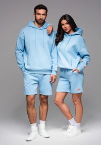 Ljusblå hoodie och shorts-set i mjukt tyg, med avslappnad passform och snörningsdetaljer, kombinerat med vita sneakers.