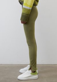 Olivgröna leggings med en slim fit, tillverkade av elastisk tyg. Har sidozippar vid anklarna. Matchas med vita sneakers med gröna detaljer.