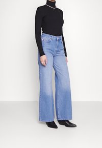 Donna che indossa jeans a gamba larga blu chiaro, dolcevita nero a maniche lunghe e stivali neri a punta che si trova di fronte a uno sfondo bianco.