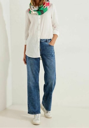 Personne portant une chemise blanche boutonnée, un jean bleu à jambes larges, des baskets blanches et une écharpe multicolore, debout avec une main dans la poche.