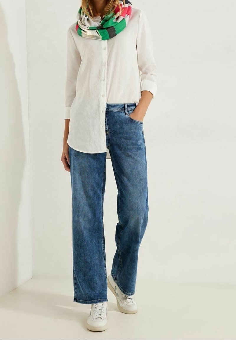 Personne portant une chemise blanche boutonnée, un jean bleu à jambes larges, des baskets blanches et une écharpe multicolore, debout avec une main dans la poche.