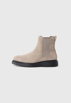 Botín Chelsea de ante beige con paneles elásticos en los laterales, un lazo para tirar y suela de goma negra. Diseño elegante con punta redondeada.