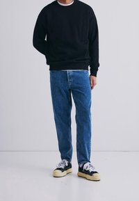 Homme portant un sweat-shirt noir, un jean bleu et des baskets noires à lacets blancs, debout devant un fond clair uni.