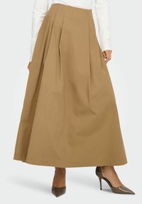 Jupe longue plissée marron en coton, avec une taille haute et une coupe fluide. Associée à des chaussures à talons hauts de couleur neutre.