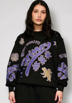 Kobieta z ciemnymi włosami do ramion nosi czarną, oversize'ową bluzę z grafiką w kolorze fioletowym i beżowym oraz wzorem z niedźwiedziem z skrzydłami.