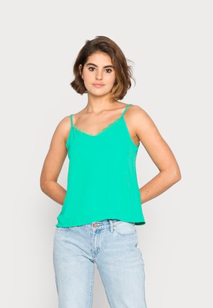 Top verde de alças finas com acabamento de renda em formato de escalope ao longo da gola, combinado com jeans de denim azul claro.