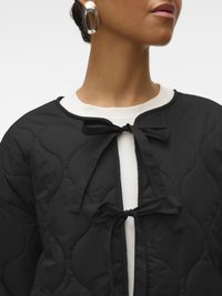 Lähikuva henkilöstä, joka käyttää mustaa tikattua takkia, joka on sidottu kahdella rusetilla valkoisen topin päällä, ja hopeisia ovaalinmuotoisia korvakoruja.