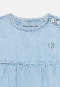 Vestido de mezclilla de color azul claro con cuello redondo, cintura fruncida y logo "CK" bordado. Dos cierres de botón en el hombro.