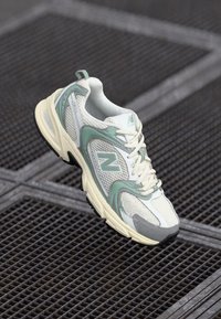 Fehér és mentazöld New Balance futócipő, amely fémsrácsok felett függ, oldalsó és felső részletességgel.